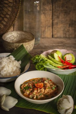 Tay Kuzey Tarzı Domuz eti ve Domates Chili Relish, ahşap masada beyaz kase içinde nam prik ong taze sebze, pişmiş pirinç, su kase ve çiçek etrafında yerleştirilen yan yemekler vardır.