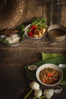 Tay Kuzey Tarzı Domuz eti ve Domates Chili Relish, ahşap masada beyaz kase içinde nam prik ong taze sebze, pişmiş pirinç, su kase ve çiçek etrafında yerleştirilen yan yemekler vardır.