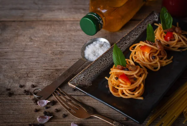 Spaghetti in Tomato sauce fit the pieces on the black plate placed on the wood table there are black pepper, fork, oil, spoon, salt and raw spaghetti placed around.