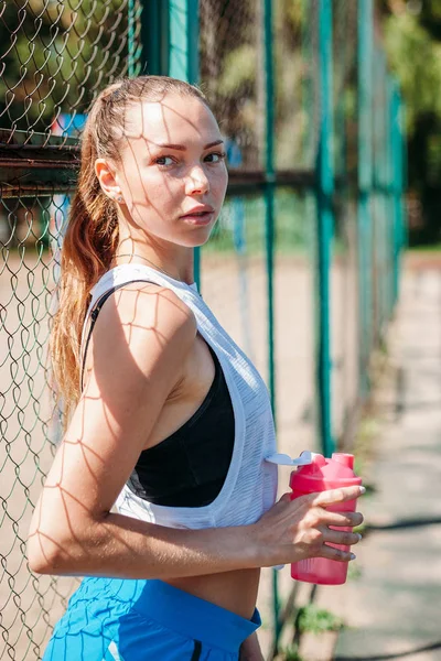 运动女孩站在墙面运动场上 手里拿着一瓶水 看着镜头 — 图库照片
