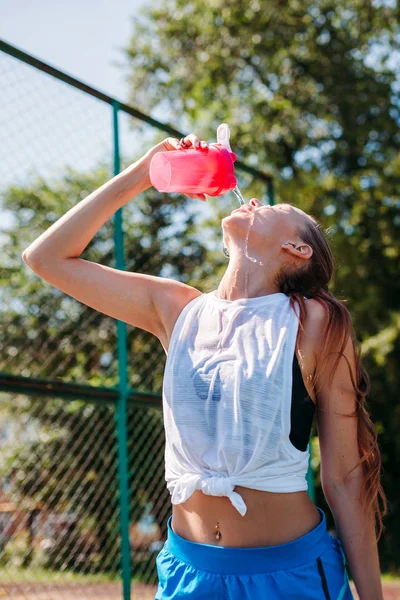 Sporty young blonde woman with a sporty bottle with cool illuminating water pours water on herself on the sports field