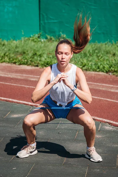 Spor yapan genç bir kadın spor lastiği bandıyla dışarıda çömeliyor.