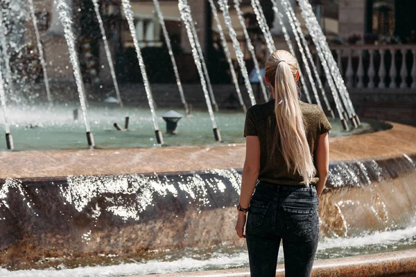 Young girl with long hair in jeans near fountain, in summer, vie