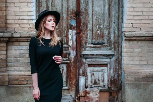 Blonde girl with long hair, in black dress in hat, stands on the — Stock Photo, Image