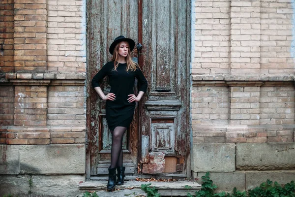 Blonde girl with long hair, in black dress in hat, stands on the — Stock Photo, Image