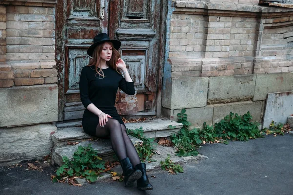 Blonde girl with long hair, in black dress in hat, is sitting on — Stock Photo, Image