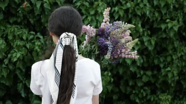 Mujer joven se para detrás con ramo de flores contra una pared verde de hojas — Vídeos de Stock