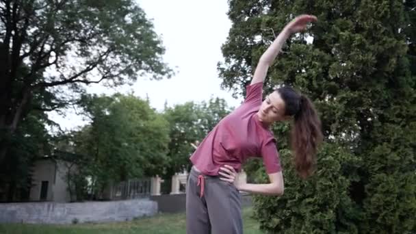 Egy fiatal barna nő tornázik a városi parkban. oldalirányú hajlítási gyakorlat — Stock videók