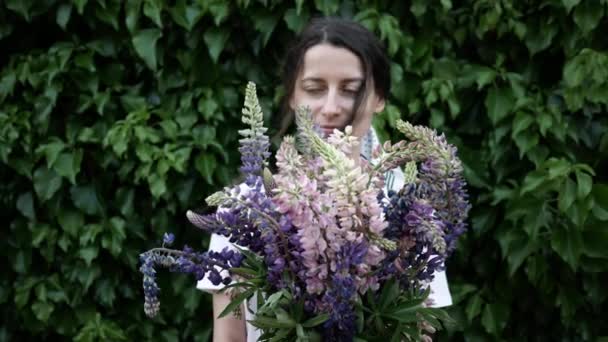 Hermosa joven con un ramo de flores cerca de una pared verde de arbustos — Vídeos de Stock