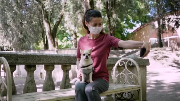 मास्क में युवा सुंदर महिला पार्क बेंच पर बैठती है और फोन और उसके कुत्ते पर सेल्फी बनाती है — स्टॉक वीडियो