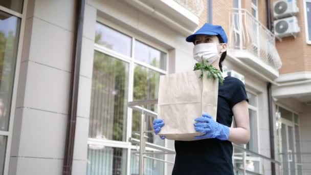 Mat leverans ung vacker kvinna i skyddsmask och handskar bär en väska med produkter från butik till hem. Livsmedelsleverans man under pandemin covid-19 — Stockvideo