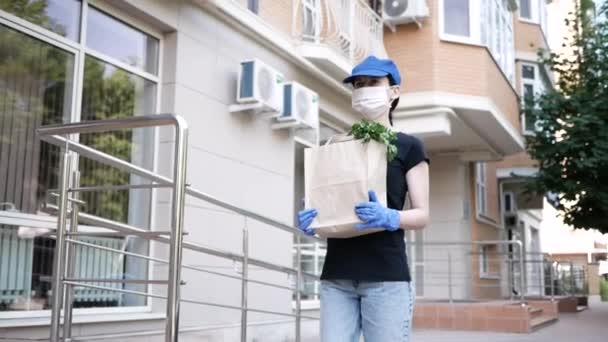 Entrega de alimentos joven hermosa mujer en máscara protectora y guantes lleva una bolsa con productos de tienda a casa. Hombre de reparto de alimentos durante la pandemia covid-19 — Vídeo de stock