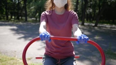 Eldivenli ve maske takan genç bir kadın Park 'taki spor aletlerinde. Halka açık yerlerde simülatör kullanımında koronavirüs salgınından korunma
