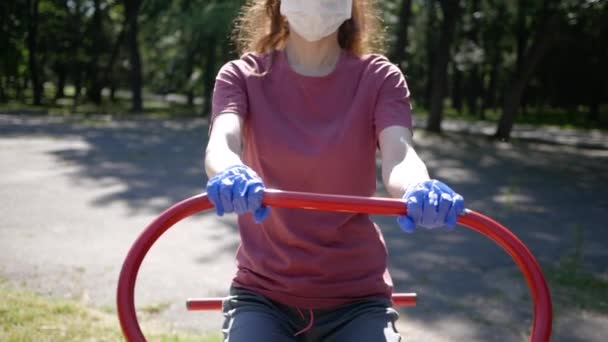 Giovane donna in guanti e maschera sportiva su attrezzature per esercizi all'aperto nel parco. Protezione da epidemia di coronavirus in uso di simulatori in luoghi pubblici — Video Stock
