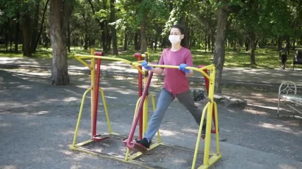 Giovane donna in maschera protettiva e guanti, entra per lo sport in attrezzature per esercizi all'aperto nel parco. Il concetto di distanza sociale — Video Stock