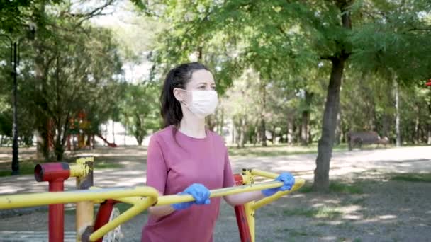 Eldivenli ve maske takan genç bir kadın Park 'taki spor aletlerinde. Halka açık yerlerde simülatör kullanımında koronavirüs salgınından korunma — Stok video