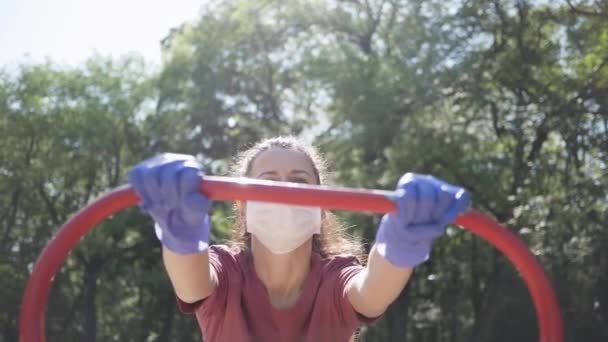 Jovem mulher em máscara protetora e luvas, vai para esportes em equipamentos de exercício ao ar livre no Parque. O conceito de distanciamento social — Vídeo de Stock