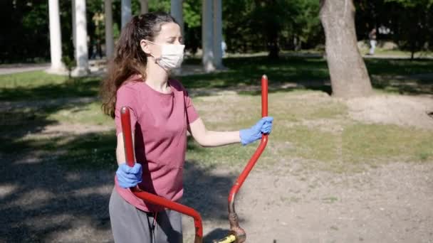 Ung kvinna i skyddande ansiktsmask och handskar, går in för sport i utomhus träningsutrustning i Park. Begreppet social distans. — Stockvideo