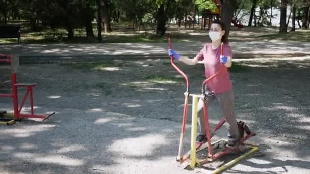 Mujer joven con mascarilla protectora y guantes, entra a practicar deportes en equipos de ejercicio al aire libre en Park. El concepto de distanciamiento social — Vídeo de stock