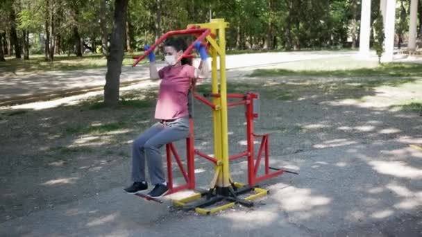 Jonge vrouw in beschermende gezichtsmasker en handschoenen, gaat in voor sport in outdoor fitnessapparatuur in Park. Het begrip "sociale afstand" — Stockvideo