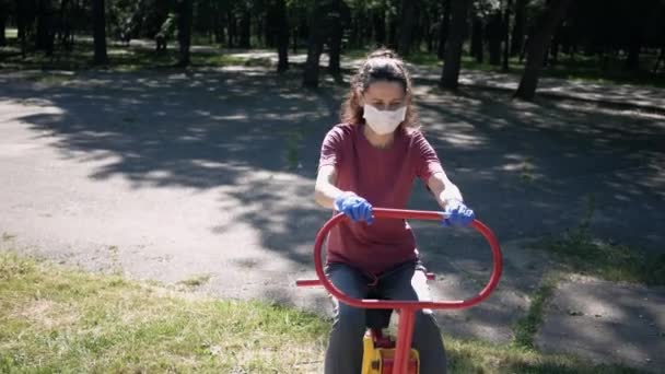 Mujer joven con mascarilla protectora y guantes, entra a practicar deportes en equipos de ejercicio al aire libre en Park. El concepto de distanciamiento social — Vídeo de stock