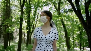 Güzel genç bir kadın yeşil yaz parkında yürüyor. Elbise ve koruyucu maske takıyor. Yürüyüşün tadını çıkarıyor.