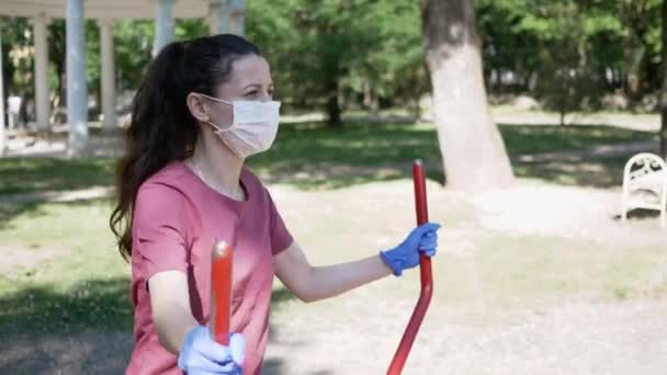 公園の屋外運動機器に手袋やフェイスマスクスポーツの若い女性。公共の場所でのシミュレーターの使用におけるコロナウイルスの流行からの保護 — ストック動画