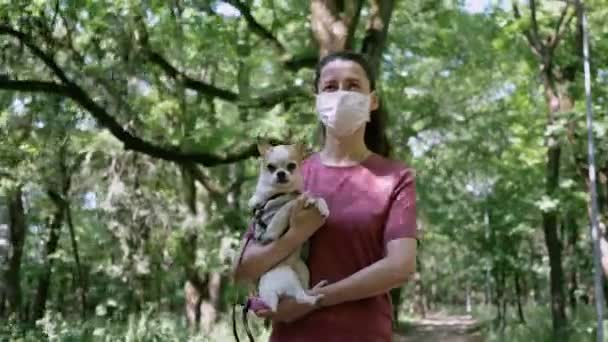 Jonge vrouw in beschermende masker wandelingen met kleine hond in Park dragen haar in de arm — Stockvideo