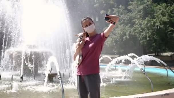 Giovane donna con maschera protettiva e piccolo cane che prende selfie alla fontana — Video Stock