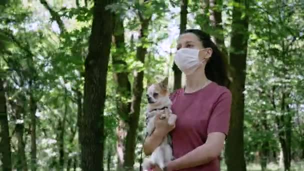 Młoda kobieta w masce ochronnej niesie jego Chihuahua na rękę w parku — Wideo stockowe