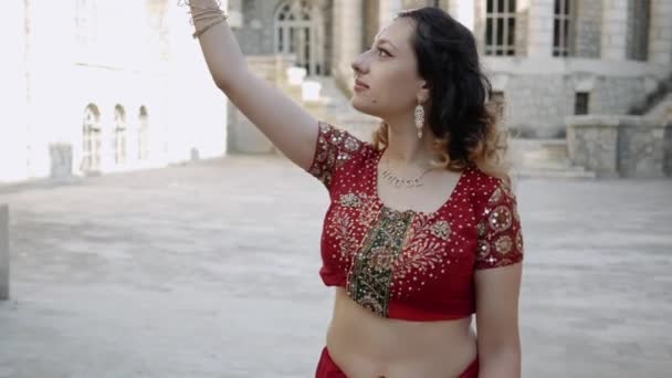 若い美しいインドの女性は、伝統的な装飾品やメフェンディのパターン、手にヘナの図面で飾られた赤いサリに身を包んだインドの古代建築の通りを歩きます — ストック動画