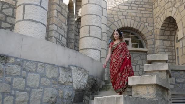 Jeune belle Indienne marche sur les rues d'architecture ancienne de la ville de l'Inde vêtue de Sari rouge, décorée avec des ornements traditionnels et des motifs Mehendi, dessins au henné sur les mains — Video