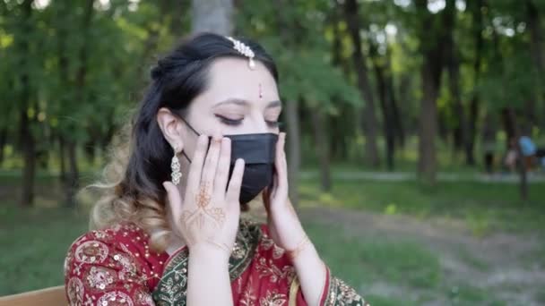 Young woman in traditional Indian outfit puts protective face mask on in Park — Stock Video