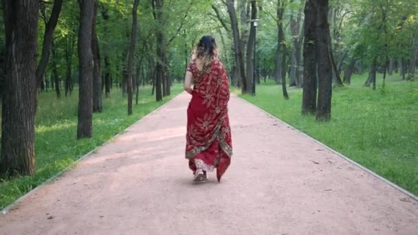 Νεαρή όμορφη γυναίκα σε ινδική εθνική κόκκινο σάρι περπάτημα στο μονοπάτι στο πάρκο — Αρχείο Βίντεο