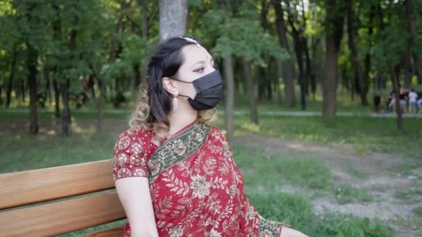 Jonge vrouw in traditionele Indiase outfit draagt beschermende gezichtsmasker op in Park — Stockvideo