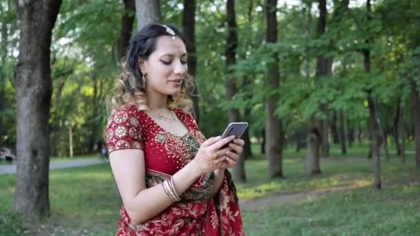 Giovane donna sorridente in rosso Sari Indiano stampe etniche SMS messaggio al telefono in estate Parco — Video Stock