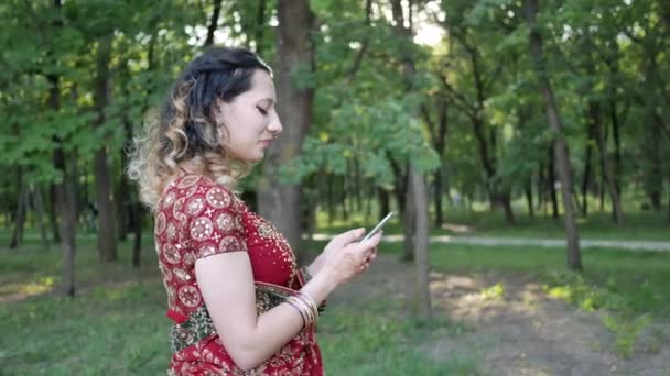 Jonge glimlachende vrouw in rood Sari Indiase etnische afdrukken SMS-bericht op de telefoon in de zomer Park — Stockvideo
