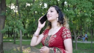 Etnik Hint kırmızısı giymiş gülümseyen genç bir kadın yazın Park 'ta telefonla konuşuyor.