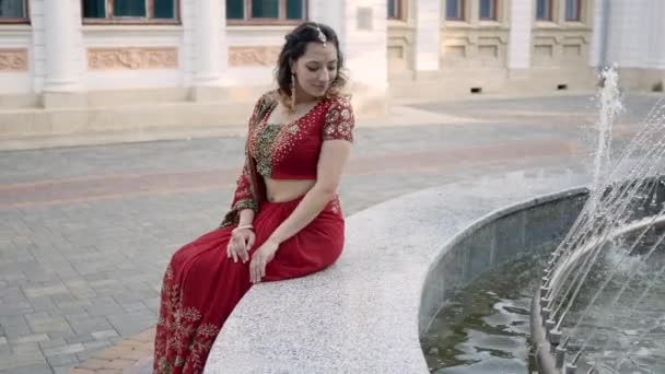 Jovem bela mulher em saris vermelhos indianos tradicionais sentado pela fonte e mão alcança o jato de fonte — Vídeo de Stock