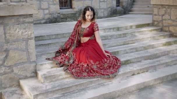 Νεαρή όμορφη Ινδή γυναίκα ποζάρουν σε δρόμους αρχαίας αρχιτεκτονικής της πόλης ντυμένοι με κόκκινο Σάρι, διακοσμημένα με παραδοσιακά στολίδια και μοτίβα Mehendi — Αρχείο Βίντεο