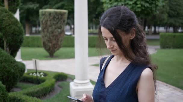 Молодая красивая брюнетка летом в парке с телефоном в руке, звонки и разговоры — стоковое видео