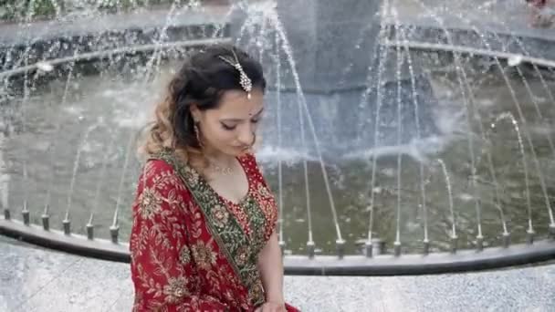 Beautiful young woman in traditional Indian red saris sitting at the fountain — Stock Video