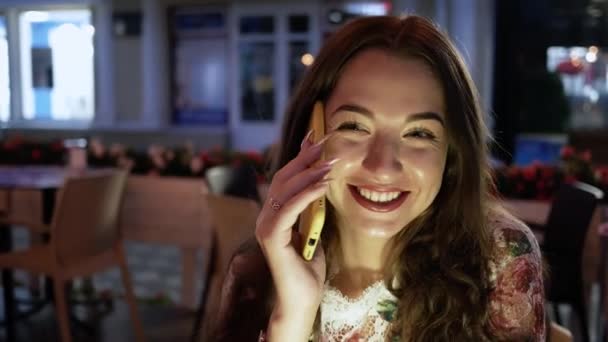 Joven hermosa mujer sentada en la noche de la mesa en la cafetería de la calle sonriendo, hablando por teléfono y riendo — Vídeo de stock