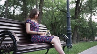 Güzel genç bir kadın, bir yaz parkında kitap okuyarak bankta oturuyor ve telefon kullanıyor.