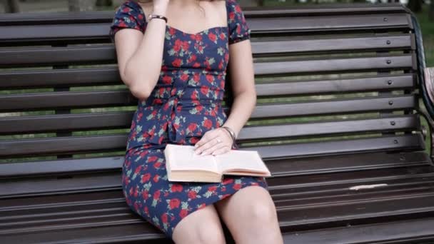 Jeune femme est assise dans le parc avec un livre, parlant au téléphone, à la recherche de quelqu'un — Video