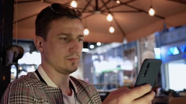 Jovem usa o telefone em um café de rua à noite, digita uma mensagem de texto ou olha através de e-mail ou redes sociais — Vídeo de Stock