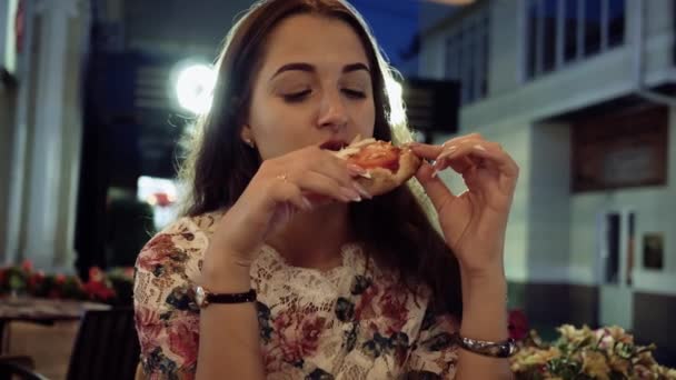 Pizzát szeletelni a kezében lány az utcai kávézóban éjszaka. Arc portré fiatal gyönyörű nő étvágya eszik szelet pizza szalámi és paradicsom — Stock videók