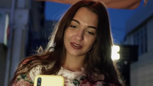 Mujer joven atractiva usando el teléfono inteligente en las calles por la noche en la cafetería — Vídeo de stock