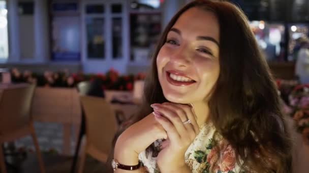 Retrato de una hermosa joven sentada en un café de moda, sonriendo y mirando a la cámara. Morena chica posando y hablando en cámara — Vídeo de stock