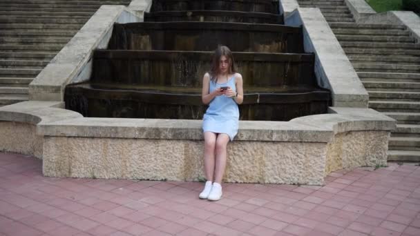 Młoda kobieta siedzi przy miejskiej fontannie i używa telefonu komórkowego. osoby uzależnione od telefonu — Wideo stockowe